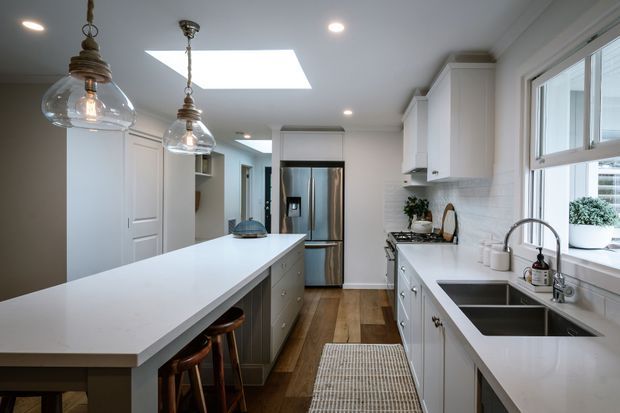 Bright, narrow galley-style kitchen with a large white island and integrated seating, white shaker cabinetry, white subway-tile backsplash, stainless-steel refrigerator, double-bowl sink beneath a window, warm timber flooring, and pendant lighting. A skylight adds natural light.
