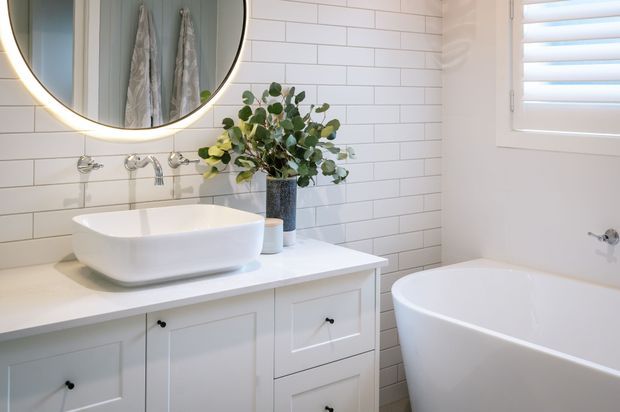 Bright, refined bathroom featuring a freestanding oval bathtub, white vessel basin on a white vanity, white subway-tile wall finish, chrome tapware, a large round illuminated mirror, and white plantation shutters. The styling is clean and contemporary with traditional panelled cabinetry.