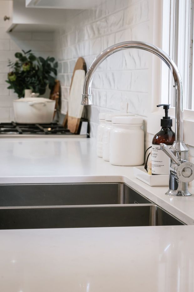 Close-up view of a bright, refined kitchen focused on a large double-bowl stainless-steel sink and tall polished chrome gooseneck faucet. White engineered-stone-looking benchtops, white textured subway-style wall tiles, white cabinetry, and white ceramic storage canisters create a clean palette. A dark glass soap dispenser and timber chopping boards add contrast, with a window providing natural light.