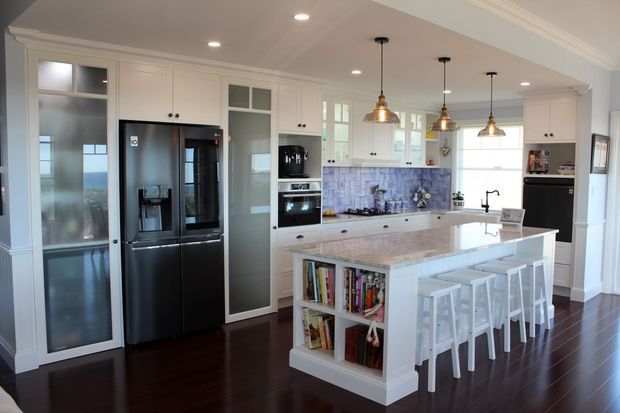 Bright, spacious white cabinetry kitchen with a large central island and seating for four. The room features dark timber flooring, a pale stone benchtop, glass-fronted pantry and display cabinetry, integrated black appliances, and a blue-toned patterned tile splashback. Pendant lights hang above the island, with coastal views visible through the glazing.