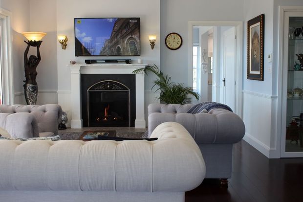 Elegant traditional living room with pale upholstered sofas arranged around a dark fireplace, wall-mounted television, decorative sconces, framed artwork, indoor plants, and dark timber flooring. White painted wall panelling and trim create a refined, formal interior.