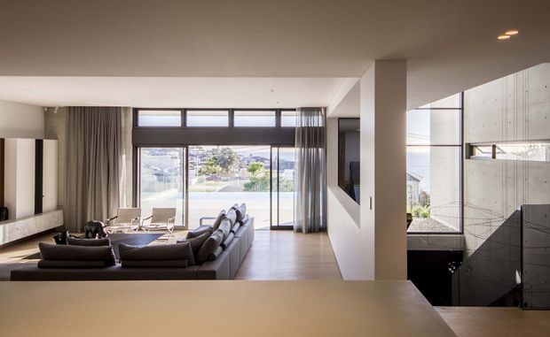 A bright contemporary open-plan living area viewed from behind a kitchen island or counter. The room features a large sectional sofa, extensive floor-to-ceiling glazing with sheer curtains, light timber flooring, a minimalist white ceiling and walls, and a prominent open stairwell with dark balustrading and concrete-toned wall surfaces. A dining area and landscaped suburban outlook are visible beyond the living room.