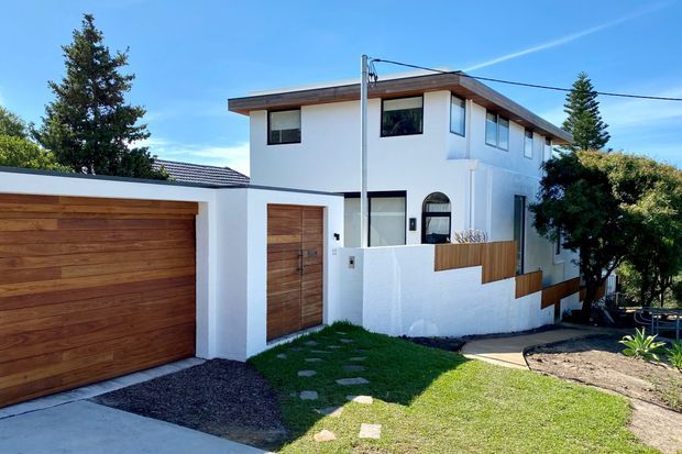 Two-storey contemporary white-rendered residence with a flat roof and projecting timber-clad roof edge. The facade features warm timber garage and entry doors, dark-framed windows, a low white boundary wall with timber accents, and a small landscaped front garden with stepping stones.