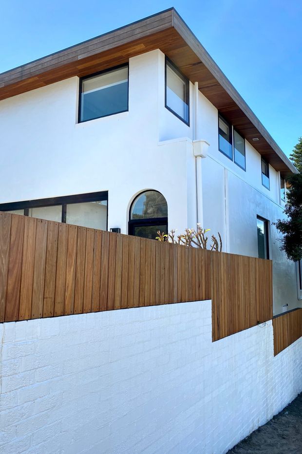 Exterior view of a contemporary two-storey residence with white rendered walls, dark-framed windows, a broad flat roof with timber soffit and fascia, and a warm vertical timber privacy fence over a white masonry boundary wall. An arched window or doorway provides a distinctive facade detail.