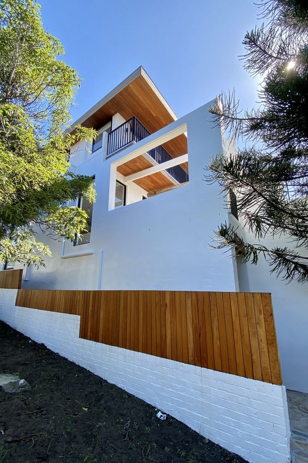 Exterior view of a contemporary multi-level residence with crisp white rendered walls, projecting balconies, dark metal balustrades, warm timber-lined soffits, and a vertical timber privacy fence set above a white-painted brick retaining wall. Mature trees frame the building.
