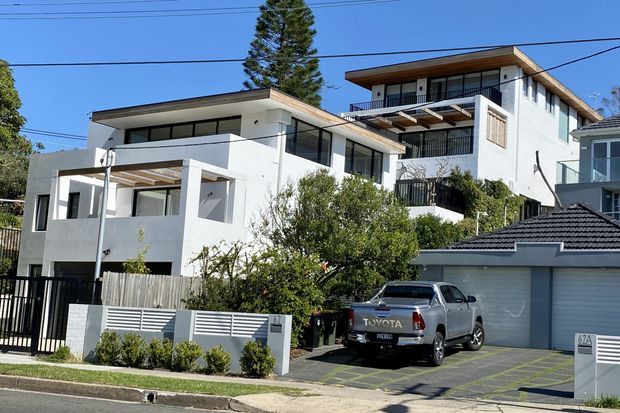 Street-level exterior view of a contemporary multi-storey residential building with white rendered walls, projecting flat rooflines, balconies, extensive glazing, black metal balustrades, and a gated driveway. Adjacent residential properties and parked vehicles are visible in the streetscape.