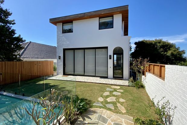 Exterior view of a compact contemporary two-storey home with a white rendered facade, broad timber-lined flat roof overhang, large ground-floor glazed openings, and an arched entry door. A landscaped front garden includes stepping-stone paving and a glass pool barrier, with timber and white masonry boundary walls.