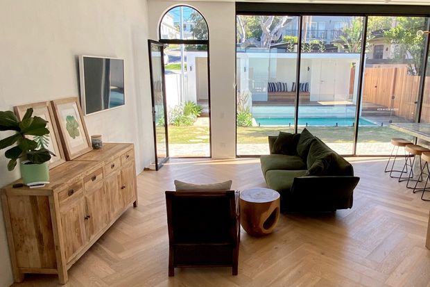 Bright contemporary living room with herringbone timber flooring, a timber sideboard, dark lounge seating, and expansive black-framed glazing opening to a landscaped courtyard and swimming pool. An arched glazed doorway adds architectural character, while a small dining or work area is visible at right.