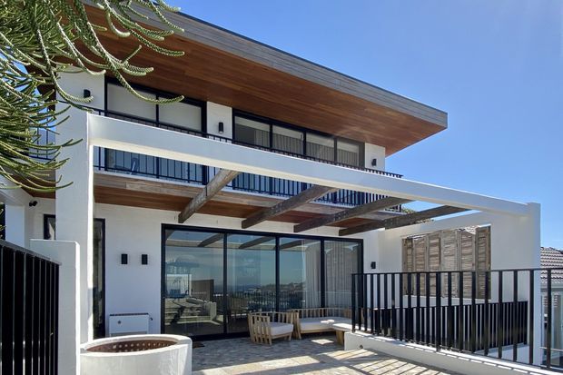 Exterior view of a contemporary multi-level residence with white rendered walls, extensive dark-framed glazing, warm timber soffits and balcony decking, black metal balustrades, and a projecting flat roof. A covered outdoor terrace with seating overlooks a distant coastal or urban horizon.