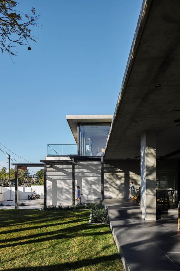 Exterior architectural photograph of a contemporary multi-level residence with broad cantilevered concrete roof planes, exposed concrete columns and soffits, extensive glazing, a glass balustrade, and dark tiled outdoor flooring. The home opens onto a landscaped lawn and covered terrace, with strong late-afternoon shadows across the facade and garden.
