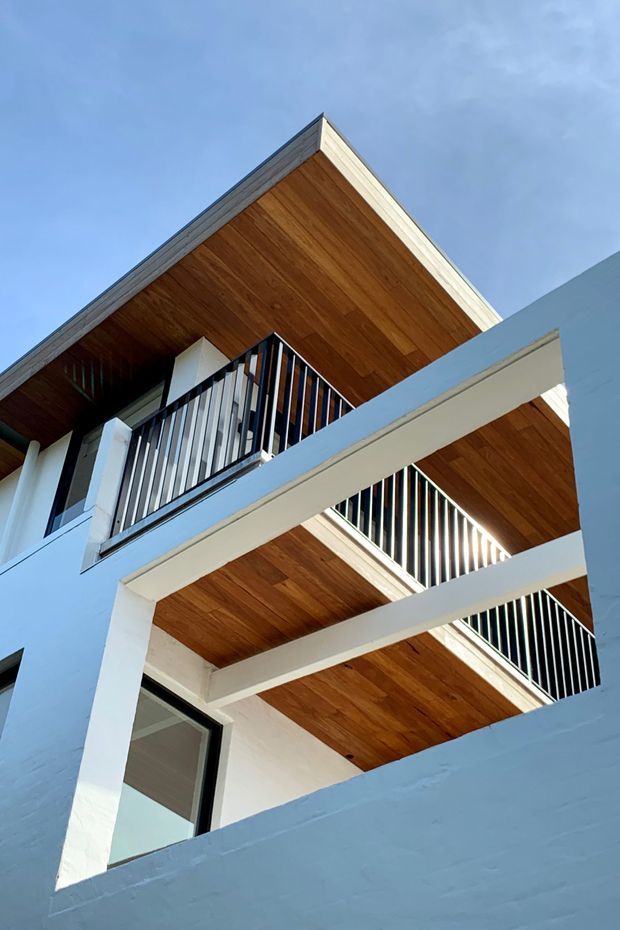 Upward-looking exterior view of a contemporary multi-level building, showing stacked balconies and terraces with white painted structural beams, dark slender metal balustrades, and warm timber-lined soffits. The facade has strong geometric lines and contrasting white, black, and wood finishes against a blue sky.