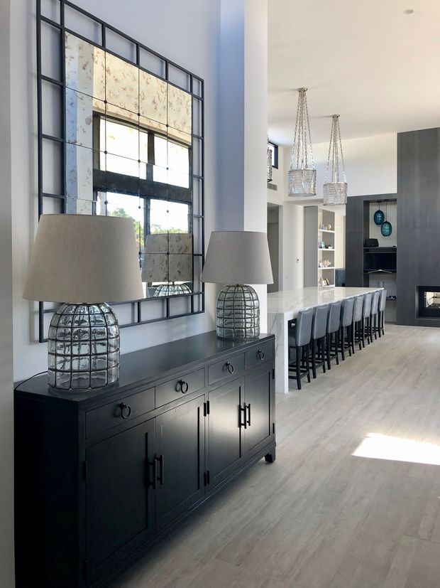 A bright, contemporary open-plan interior featuring a black sideboard with glass-and-metal table lamps beneath a large industrial-style grid mirror. Beyond it is a kitchen with a long island, upholstered bar stools, pendant lights, white cabinetry, dark wall joinery, and pale timber-look flooring.