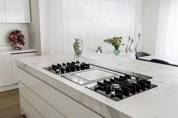 Bright, refined contemporary kitchen dominated by a large white marble-look island with integrated black gas cooktops and a flush-mounted sink. White handleless cabinetry, sheer floor-to-ceiling curtains, pale timber flooring, metallic decorative vessels, and black dining chairs create a clean, luxurious monochromatic interior.