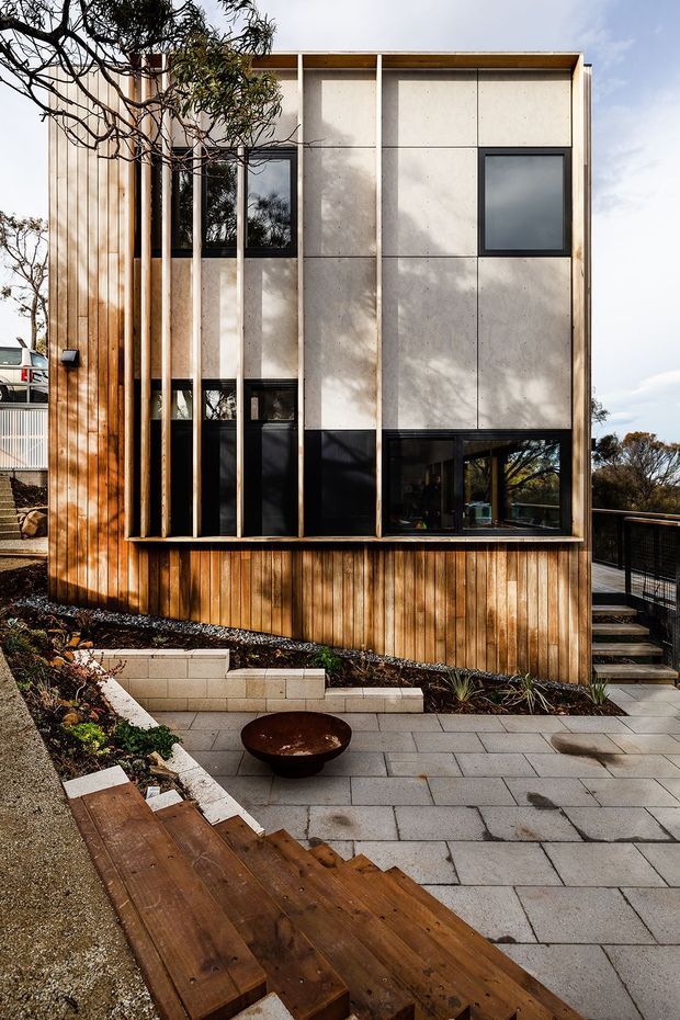 Exterior view of a compact contemporary multi-storey residence on a sloped site. The facade combines vertical warm timber cladding with large pale concrete panels, dark-framed glazing, and projecting vertical timber fins. A paved terrace, timber steps, retaining walls, planted garden beds, and a dark metal balustrade surround the building.