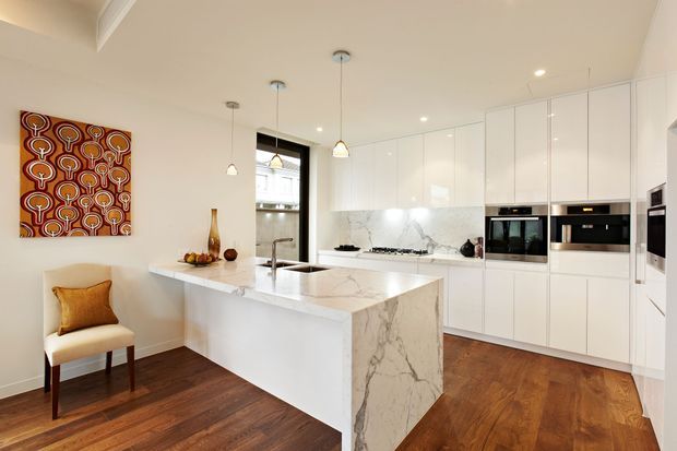 Bright, refined white kitchen with a large marble island, extensive glossy white cabinetry, integrated appliances, pendant lighting and warm dark timber flooring. A patterned artwork and upholstered chair add colour and character.