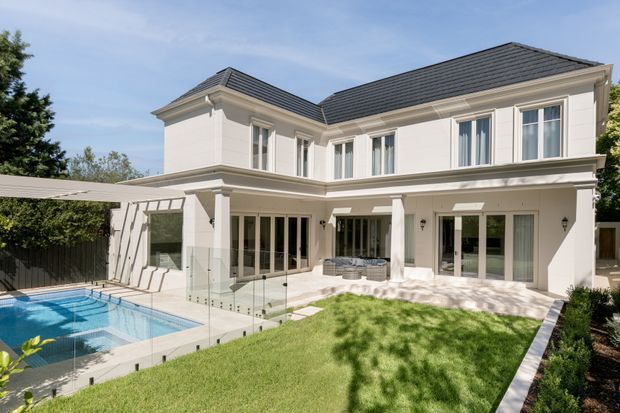 A large two-storey white rendered residence with a symmetrical, classically influenced facade and dark grey tiled hipped roofs. The rear of the house opens to a landscaped lawn, paved terrace, covered outdoor seating area, and a blue swimming pool enclosed by frameless glass safety fencing. Tall multi-pane windows and French doors provide extensive glazing, with white columns and decorative cornices adding formal detailing.