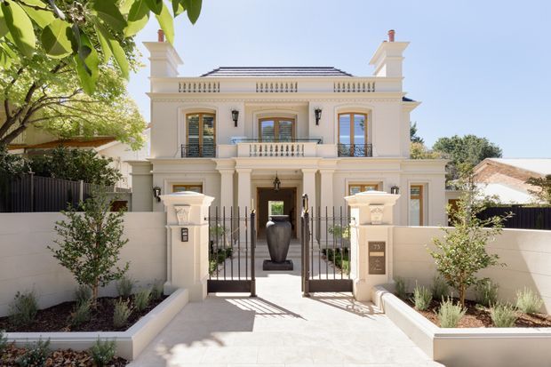 Front exterior view of a grand two-storey residence with a symmetrical cream-rendered facade, classical columns and pilasters, ornate balustrades, decorative cornices, shuttered windows, and a central gated entry. Low landscaped garden beds and rendered boundary walls frame the pale paved approach.