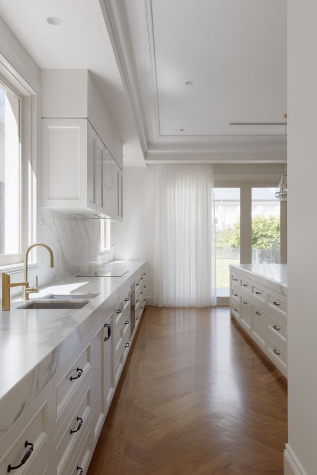 Bright, refined galley-style kitchen with extensive white shaker cabinetry, veined marble-look benchtops and splashback, brass tapware, dark hardware, and a central island. The room features a warm herringbone timber floor, coffered and panelled ceiling detailing, recessed lighting, full-height windows with sheer curtains, and glazed doors opening toward greenery.