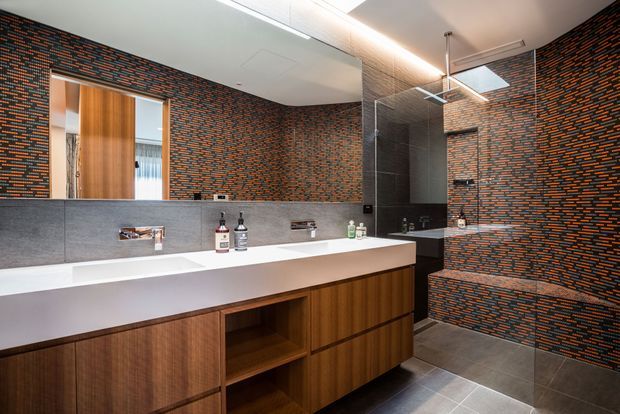Contemporary bathroom with a long double vanity featuring a white integrated benchtop and warm timber cabinetry. Large mirrors reflect the room, while the walls and shower area are finished in a striking dark mosaic tile with orange and blue accents. A walk-in shower, wall-mounted fixtures, grey tiled floor, and linear ceiling lighting complete the refined interior.