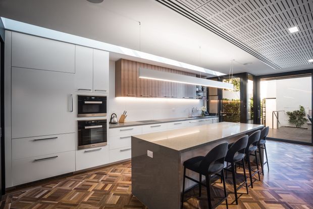 A spacious contemporary kitchen with a long central island and integrated bar seating for four. The room features full-height white cabinetry, built-in double ovens, a pale stone benchtop, a timber-toned feature cabinet with open shelving and under-cabinet lighting, dark-framed glazing, and a geometric parquet timber floor. A linear slatted ceiling and abundant daylight contribute to the polished, open-plan feel.