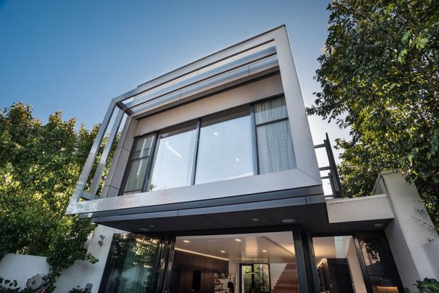 Low-angle exterior view of a contemporary multi-level house with a crisp white and dark grey facade, extensive floor-to-ceiling glazing, projecting upper storey, and horizontal roof-edge screening. Trees frame the building, while the illuminated open-plan interior and staircase are visible through the ground-floor glazing.