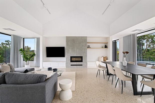 A bright, open-plan contemporary living and dining area with floor-to-ceiling glazing on multiple sides. The room features a grey sectional sofa, minimalist coffee table and side table, a large dark dining table with upholstered chairs, a wall-mounted television, built-in shelving, and a simple fireplace set within a light concrete-look feature wall. Pale terrazzo-like flooring, white walls, sheer curtains, and indoor greenery create an airy, refined interior.