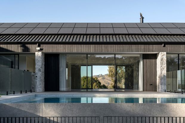 Contemporary single-storey residence viewed from the swimming pool, featuring a long low roofline, dark vertical timber cladding, extensive floor-to-ceiling glazing, and pale natural-stone columns. The reflective pool occupies the foreground, while a dry hilly landscape is visible through the central glazing. The facade has a restrained, linear composition with a dark tiled roof and prominent covered outdoor edge.