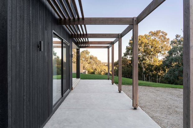 A contemporary outdoor terrace runs alongside a dark vertically clad building, framed by a timber pergola with projecting rafters and substantial square posts. Large glazed sliding doors open onto the pale concrete terrace, which overlooks a gently sloping grassy and wooded landscape.