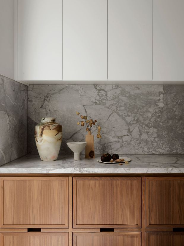 Close-up view of a refined built-in kitchen or storage joinery installation. Warm timber cabinetry with recessed panel doors sits beneath a pale grey veined stone benchtop and full-height matching splashback. Minimal white overhead cupboards create a clean, contemporary composition, with a few decorative vessels and fruit arranged on the counter.