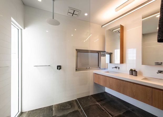 Bright contemporary bathroom featuring a long floating timber vanity with a pale benchtop, integrated twin basins, large mirrors, linear LED lighting, white wall tiling, dark large-format floor tiles, a frameless glass shower enclosure, and a high horizontal window.