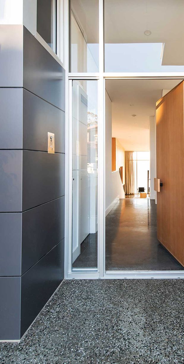 A modern residential entrance viewed from outside, with dark charcoal panel cladding, a full-height glazed entry surround, a large timber pivot door, and a polished aggregate or terrazzo-like threshold. The bright hallway beyond has a dark polished floor, white walls, timber joinery, and strong natural light from a skylight and rear glazing.
