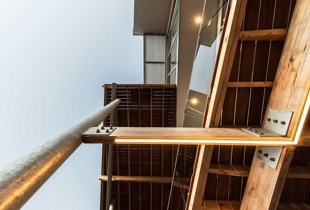 Upward-looking architectural view beneath a timber-framed canopy or roof structure. Exposed warm timber beams, dark timber slats, metal connection plates, a cylindrical metal post, and reflective glazing create a strong structural composition with integrated warm lighting.