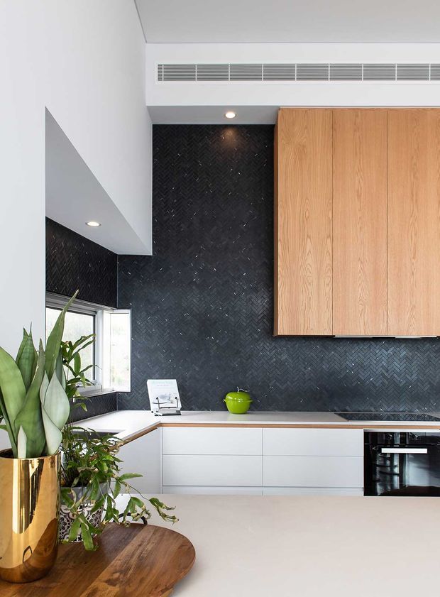 A contemporary kitchen with white lower cabinetry, a large natural timber overhead cabinet bank, and a dark charcoal herringbone tile backsplash. The kitchen includes a pale benchtop, integrated oven, stainless ventilation grille, recessed downlights, and a window providing natural light. Large leafy plants in a metallic gold planter and a wooden table occupy the foreground.