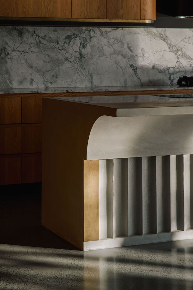 Close-up interior view of a sophisticated kitchen featuring dark timber cabinetry, a dramatic grey-veined stone splashback and benchtop, and a sculptural island with a curved light-toned counter edge and vertically fluted front panel. The polished concrete floor reflects strong directional light, creating a moody, high-contrast atmosphere.
