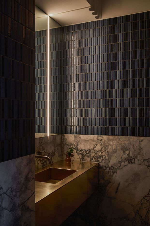 A dark, luxurious bathroom vanity viewed in a narrow space. Deep blue vertically arranged wall tiles cover the upper walls, while dramatic veined marble forms the lower wall and backsplash surfaces. A sculptural bronze-gold vanity with an integrated basin sits beneath a mirror, accented by warm concealed lighting and a small decorative vase. The atmosphere is moody and refined.