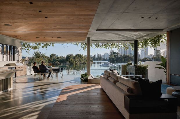 A spacious, open-plan contemporary living area with a kitchen and dining zone opening directly onto a waterfront terrace. Floor-to-ceiling glazing frames a calm body of water, tree-covered shoreline, and distant high-rise buildings. The room features warm timber ceiling and flooring, a polished concrete ceiling section, slim dark structural columns, a neutral sectional sofa, and abundant natural light.