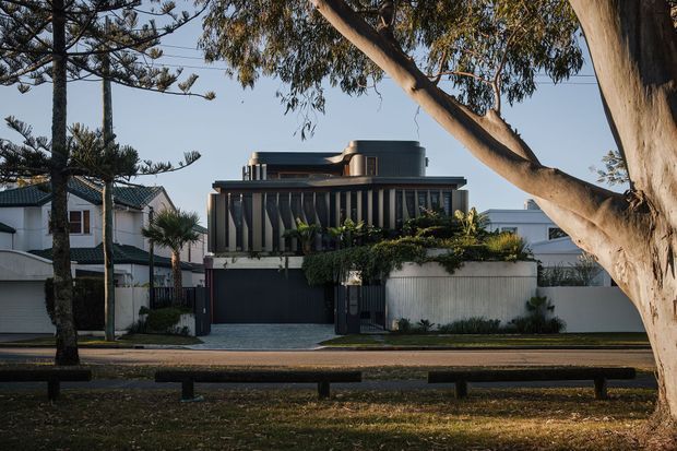 Street-facing view of a contemporary multi-storey residence with a dark vertical-slat facade, flat roof, rooftop cylindrical feature, garage entry, and a pale rendered boundary wall. Mature trees and neighboring homes frame the suburban setting.