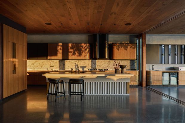 A sophisticated open-plan kitchen with a large island, integrated timber cabinetry, marble or natural-stone splashback, and a distinctive ribbed island front. The space has a timber-lined ceiling, polished dark flooring, black-framed glazing or sliding doors, and warm low-angle sunlight. A secondary kitchen or utility area is visible beyond the opening on the right.