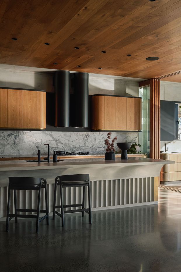 A sophisticated contemporary kitchen with warm timber ceiling and cabinetry, a dramatic veined stone splashback, dark cylindrical rangehoods, and a large island with a sculptural fluted base. Two black bar stools sit at the island, while vertical timber screening and glazing connect the kitchen to an adjacent space. The palette is warm timber, pale stone, charcoal, and dark grey, with polished reflective flooring and restrained decorative objects.