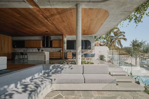 A sophisticated open-plan indoor-outdoor living area with a large light-grey sectional sofa in the foreground, a timber-lined ceiling with curved detailing, exposed concrete slab and column, and a contemporary kitchen with dark timber cabinetry and a stone island. Frameless glass balustrades overlook a swimming pool and tropical landscaping, creating a relaxed resort-like setting.