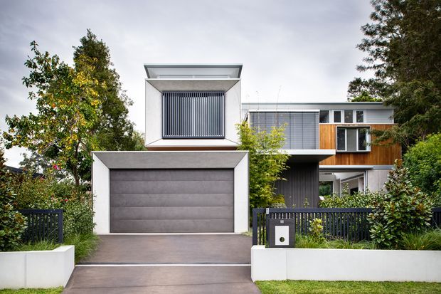 Street-facing photograph of a contemporary multi-level residence with a prominent integrated double garage, flat rooflines, projecting box forms, vertical screening, large windows, dark metal fencing, white rendered walls and landscaped planting. The house sits in a leafy suburban setting.