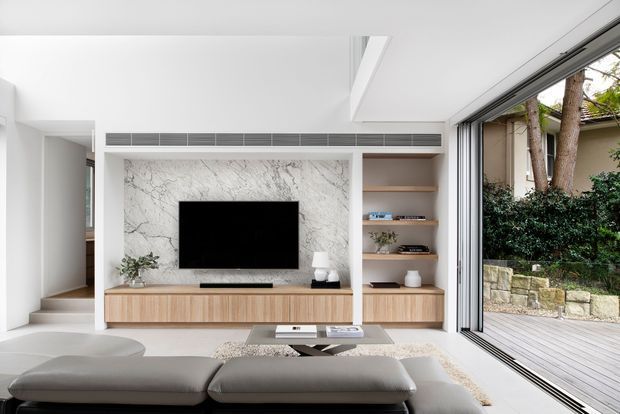 Bright contemporary living room with a low-profile grey sectional sofa, pale timber built-in entertainment joinery, open shelving, and a large marble-look feature wall surrounding the television. Floor-to-ceiling sliding glass doors open to a timber deck and landscaped exterior, while a skylight or high-level ceiling opening adds natural light.