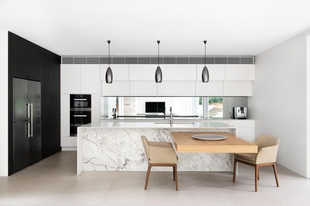 Bright contemporary kitchen with a large marble-look waterfall island and integrated dining table. The room features extensive white flat-panel cabinetry, a black full-height wall with integrated appliances and refrigerator, dark pendant lights, pale flooring, and a linear ventilation grille above the cabinetry. Two dining chairs are positioned at the timber table extension, with a window and sink area behind.