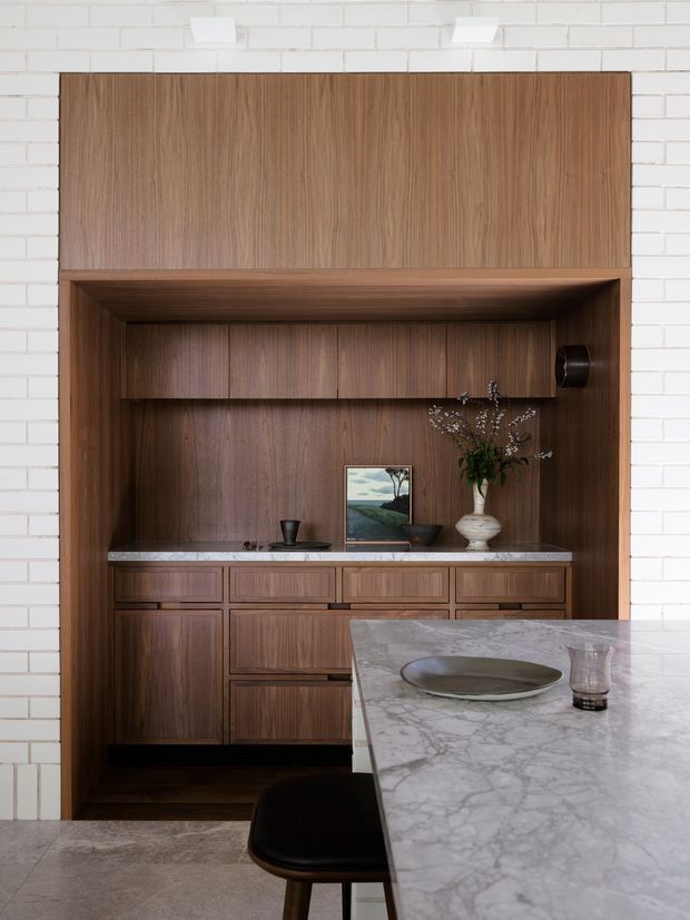 A refined kitchen interior featuring a built-in walnut-toned timber cabinetry wall set within a white painted brick surround. The cabinetry includes a recessed display and work area with drawers, cupboards, stone benchtop, shelving, decorative objects and a vase. In the foreground, a large pale veined stone island with dark seating creates a strong focal point. The overall composition is warm, tailored and contemporary with subtle mid-century influences.