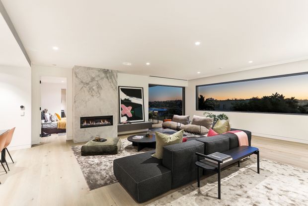 Contemporary open-plan living room with a large dark sectional sofa, layered rugs, low coffee tables, a marble-look fireplace feature wall, recessed lighting, and wide horizontal glazing overlooking the landscape at dusk. A bedroom is visible through the open doorway in the background.
