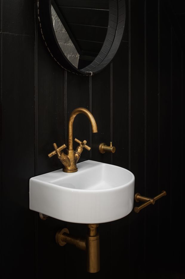A dramatic close-up of a compact bathroom washbasin against black vertical wall panelling. The white wall-mounted basin is paired with aged brass/gold exposed plumbing, a curved gooseneck tap, cross-handle fittings, and a round black-framed mirror. The dark palette and metallic fixtures create a refined, moody, high-contrast interior.