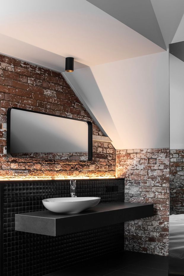 A dramatic contemporary bathroom interior with an exposed, distressed brick wall, black square-textured wall tiles, a floating matte-black vanity, and a white vessel basin. A large rounded-rectangle mirror, wall-mounted black pendant light, concealed strip lighting, and angular white ceiling forms create a high-contrast industrial aesthetic. A partial bathtub or shower area is visible at the right edge.