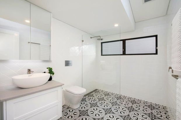 Bright contemporary bathroom with white tiled walls, a frameless glass shower enclosure, wall-mounted toilet, floating white vanity with a round vessel basin, mirrored cabinetry, and patterned black-and-white floor tiles. A narrow high window provides natural light.