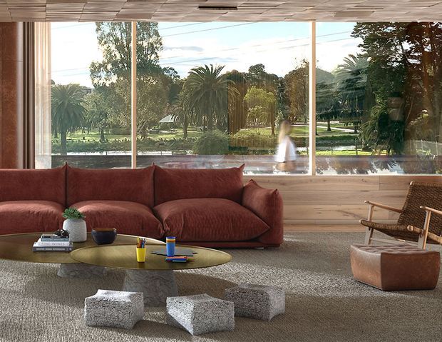 Warm, contemporary living room with a large rust-coloured sectional sofa facing a low oval coffee table. Full-height windows overlook a landscaped garden, while a timber-clad lower wall and distinctive timber ceiling create a natural, textured interior. A woven lounge chair, leather ottoman, textured rug and sculptural stone-like stools complete the relaxed setting.