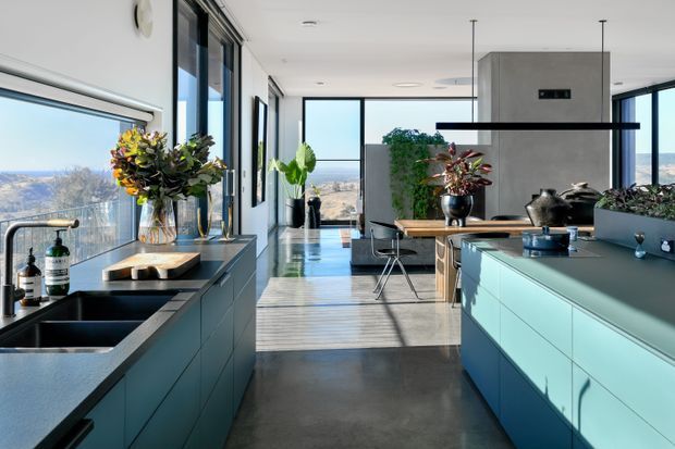 A bright, open-plan contemporary kitchen with a long central island and extensive floor-to-ceiling glazing overlooking a dry rural landscape. The kitchen features streamlined blue-grey cabinetry, dark stone benchtops, integrated appliances, a large black sink, pendant lighting, indoor plants, and a dining area beyond. Strong natural light and polished concrete flooring create an airy, refined atmosphere.